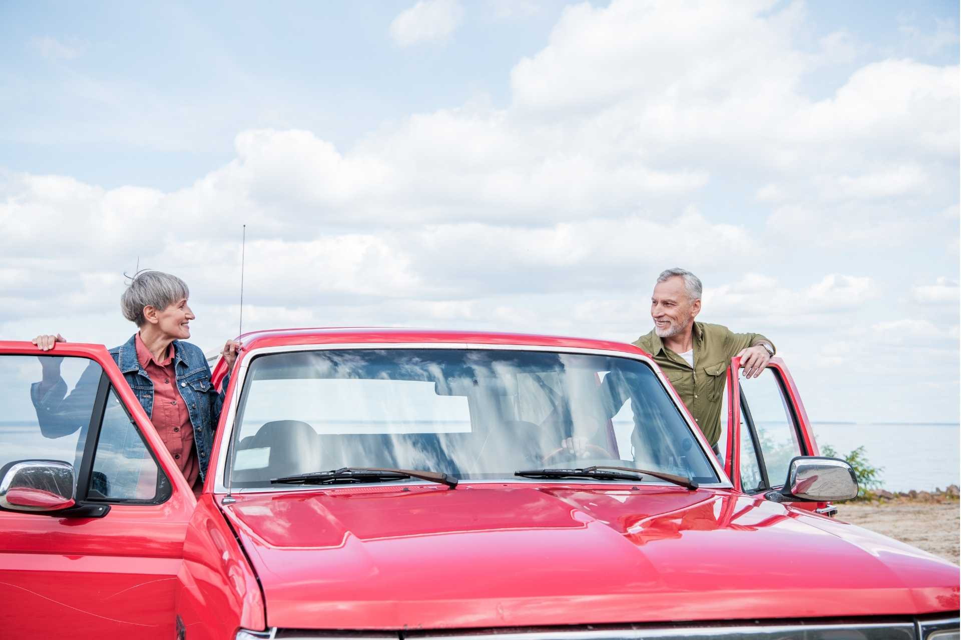 A senior couple getting into a red vintage car and smiling at each other.