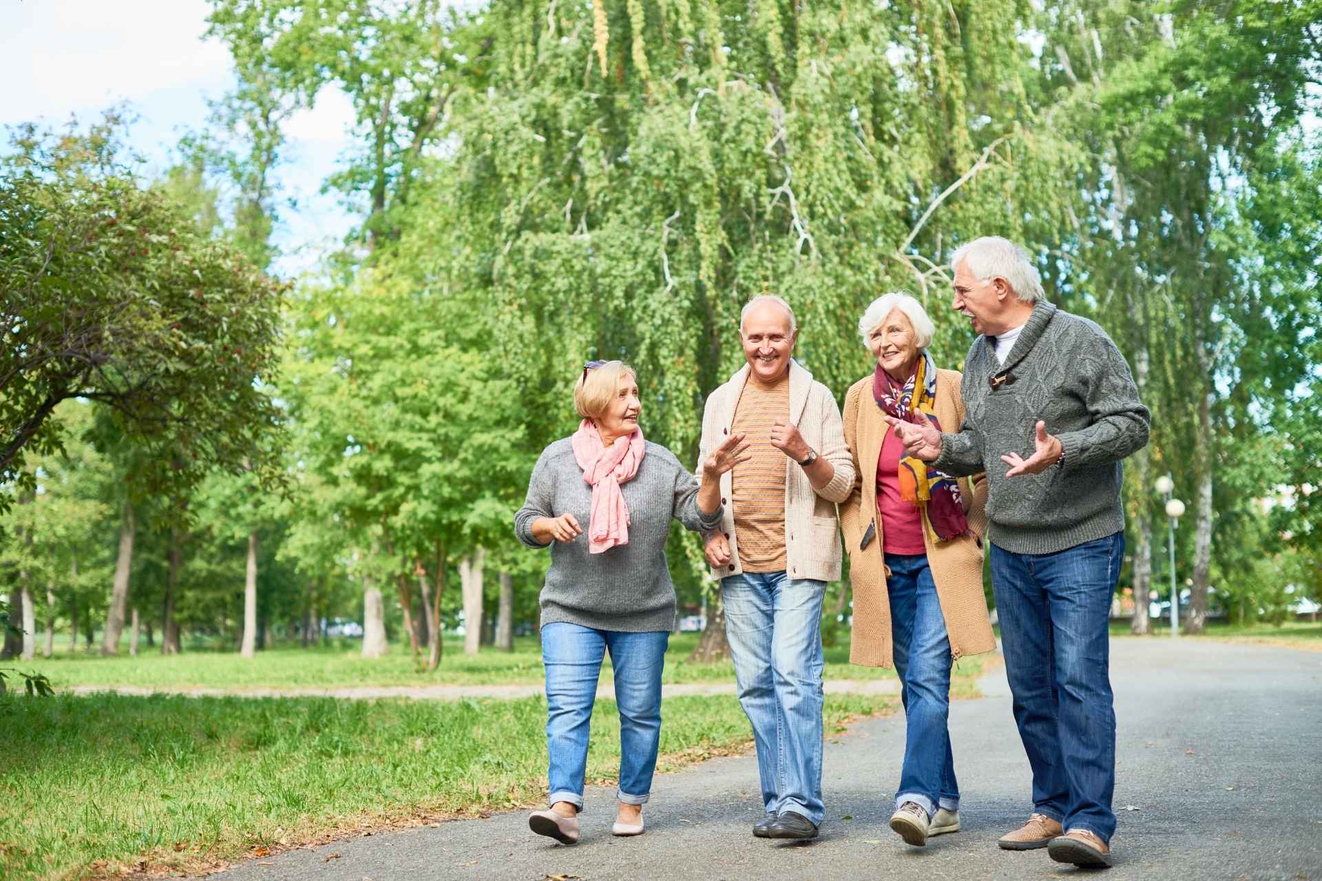 Four older adults walking together on a paved path in a green park, smiling and enjoying conversation during a leisurely outdoor stroll.